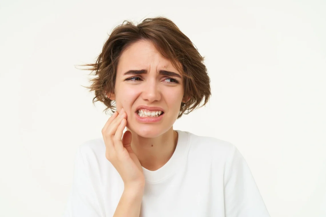 A young woman winces and clutches her jaw in severe discomfort from wisdom tooth pain.
